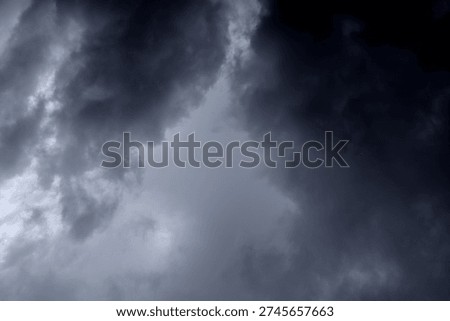 The image captures a dramatic expanse of sky dominated by towering storm clouds. Thick, heavy masses of dark gray and charcoal clouds loom from both sides of the frame, curving inward as if closing around a brighter opening at the center. The clouds appear dense and textured, with soft, billowing edges that suggest constant movement and turbulence. Their forms are layered and sculpted, with deeper shadows pooling in the folds and lighter grays tracing the contours where faint light filters through.
At the center of the composition, a pale, diffused glow breaks through the gloom. This lighter patch of sky, almost silvery in tone, creates a striking contrast against the surrounding darkness. It feels like a temporary clearing, a narrow corridor of light between two advancing walls of cloud. The brightness does not come from direct sunlight but rather from sunlight obscured by thick vapor, giving the scene a moody, atmospheric quality.
The left side of the image shows clouds with slightly more variation in tone, where subtle gradations of gray transition from smoky light to near-black. The right side appears heavier and darker, with a more imposing, shadowed presence. The cloud formations rise upward and inward, giving the impression of height and scale, as though the viewer is looking up from the ground at a vast, shifting ceiling of weather.
There are no visible landmarks, buildings, or horizon lines—only sky. This absence of grounding elements enhances the feeling of immersion, as if the viewer is enveloped by the storm. The image conveys tension and anticipation, suggesting that rain or thunder may soon follow. At the same time, the central glow hints at resilience and contrast—light pushing gently against encroaching darkness. The overall mood is powerful, contemplative, and slightly ominous, capturing the raw, ever-changing drama of the atmosphere.