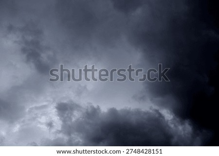 The image captures a dramatic expanse of sky dominated by towering storm clouds. Thick, heavy masses of dark gray and charcoal clouds loom from both sides of the frame, curving inward as if closing around a brighter opening at the center. The clouds appear dense and textured, with soft, billowing edges that suggest constant movement and turbulence. Their forms are layered and sculpted, with deeper shadows pooling in the folds and lighter grays tracing the contours where faint light filters through.
At the center of the composition, a pale, diffused glow breaks through the gloom. This lighter patch of sky, almost silvery in tone, creates a striking contrast against the surrounding darkness. It feels like a temporary clearing, a narrow corridor of light between two advancing walls of cloud. The brightness does not come from direct sunlight but rather from sunlight obscured by thick vapor, giving the scene a moody, atmospheric quality.
The left side of the image shows clouds with slightly more variation in tone, where subtle gradations of gray transition from smoky light to near-black. The right side appears heavier and darker, with a more imposing, shadowed presence. The cloud formations rise upward and inward, giving the impression of height and scale, as though the viewer is looking up from the ground at a vast, shifting ceiling of weather.
There are no visible landmarks, buildings, or horizon lines—only sky. This absence of grounding elements enhances the feeling of immersion, as if the viewer is enveloped by the storm. The image conveys tension and anticipation, suggesting that rain or thunder may soon follow. At the same time, the central glow hints at resilience and contrast—light pushing gently against encroaching darkness. The overall mood is powerful, contemplative, and slightly ominous, capturing the raw, ever-changing drama of the atmosphere.