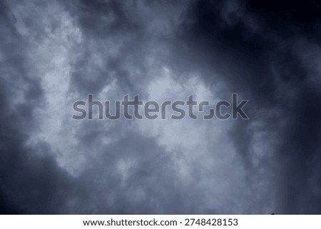 The image captures a dramatic expanse of sky dominated by towering storm clouds. Thick, heavy masses of dark gray and charcoal clouds loom from both sides of the frame, curving inward as if closing around a brighter opening at the center. The clouds appear dense and textured, with soft, billowing edges that suggest constant movement and turbulence. Their forms are layered and sculpted, with deeper shadows pooling in the folds and lighter grays tracing the contours where faint light filters through.
At the center of the composition, a pale, diffused glow breaks through the gloom. This lighter patch of sky, almost silvery in tone, creates a striking contrast against the surrounding darkness. It feels like a temporary clearing, a narrow corridor of light between two advancing walls of cloud. The brightness does not come from direct sunlight but rather from sunlight obscured by thick vapor, giving the scene a moody, atmospheric quality.
The left side of the image shows clouds with slightly more variation in tone, where subtle gradations of gray transition from smoky light to near-black. The right side appears heavier and darker, with a more imposing, shadowed presence. The cloud formations rise upward and inward, giving the impression of height and scale, as though the viewer is looking up from the ground at a vast, shifting ceiling of weather.
There are no visible landmarks, buildings, or horizon lines—only sky. This absence of grounding elements enhances the feeling of immersion, as if the viewer is enveloped by the storm. The image conveys tension and anticipation, suggesting that rain or thunder may soon follow. At the same time, the central glow hints at resilience and contrast—light pushing gently against encroaching darkness. The overall mood is powerful, contemplative, and slightly ominous, capturing the raw, ever-changing drama of the atmosphere.