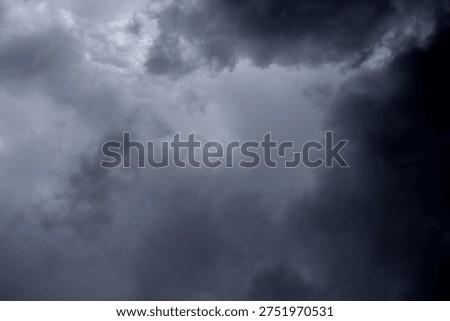 The image captures a dramatic expanse of sky dominated by towering storm clouds. Thick, heavy masses of dark gray and charcoal clouds loom from both sides of the frame, curving inward as if closing around a brighter opening at the center. The clouds appear dense and textured, with soft, billowing edges that suggest constant movement and turbulence. Their forms are layered and sculpted, with deeper shadows pooling in the folds and lighter grays tracing the contours where faint light filters through.
At the center of the composition, a pale, diffused glow breaks through the gloom. This lighter patch of sky, almost silvery in tone, creates a striking contrast against the surrounding darkness. It feels like a temporary clearing, a narrow corridor of light between two advancing walls of cloud. The brightness does not come from direct sunlight but rather from sunlight obscured by thick vapor, giving the scene a moody, atmospheric quality.
The left side of the image shows clouds with slightly more variation in tone, where subtle gradations of gray transition from smoky light to near-black. The right side appears heavier and darker, with a more imposing, shadowed presence. The cloud formations rise upward and inward, giving the impression of height and scale, as though the viewer is looking up from the ground at a vast, shifting ceiling of weather.
There are no visible landmarks, buildings, or horizon lines—only sky. This absence of grounding elements enhances the feeling of immersion, as if the viewer is enveloped by the storm. The image conveys tension and anticipation, suggesting that rain or thunder may soon follow. At the same time, the central glow hints at resilience and contrast—light pushing gently against encroaching darkness. The overall mood is powerful, contemplative, and slightly ominous, capturing the raw, ever-changing drama of the atmosphere.