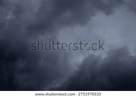 The image captures a dramatic expanse of sky dominated by towering storm clouds. Thick, heavy masses of dark gray and charcoal clouds loom from both sides of the frame, curving inward as if closing around a brighter opening at the center. The clouds appear dense and textured, with soft, billowing edges that suggest constant movement and turbulence. Their forms are layered and sculpted, with deeper shadows pooling in the folds and lighter grays tracing the contours where faint light filters through.
At the center of the composition, a pale, diffused glow breaks through the gloom. This lighter patch of sky, almost silvery in tone, creates a striking contrast against the surrounding darkness. It feels like a temporary clearing, a narrow corridor of light between two advancing walls of cloud. The brightness does not come from direct sunlight but rather from sunlight obscured by thick vapor, giving the scene a moody, atmospheric quality.
The left side of the image shows clouds with slightly more variation in tone, where subtle gradations of gray transition from smoky light to near-black. The right side appears heavier and darker, with a more imposing, shadowed presence. The cloud formations rise upward and inward, giving the impression of height and scale, as though the viewer is looking up from the ground at a vast, shifting ceiling of weather.
There are no visible landmarks, buildings, or horizon lines—only sky. This absence of grounding elements enhances the feeling of immersion, as if the viewer is enveloped by the storm. The image conveys tension and anticipation, suggesting that rain or thunder may soon follow. At the same time, the central glow hints at resilience and contrast—light pushing gently against encroaching darkness. The overall mood is powerful, contemplative, and slightly ominous, capturing the raw, ever-changing drama of the atmosphere.