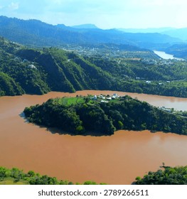 The image captures an elevated, high-angle panoramic view of a sweeping river valley, featuring a prominent green island or peninsula at its center.
​Central Focal Point
​The Island: A large, elevated mound of land sits in the middle of the river, heavily covered in dark green trees and vegetation.
​Structures: On the flatter, upper ridge of this island, there is a small cluster of white buildings, including one with a distinct white dome, suggesting a religious site, shrine, or small settlement. Adjacent to these buildings are small, bright green patches of cultivated fields.
​River and Waterway
​The Water: The river wraps around the central landmass in a wide bend. The water has a distinct, opaque brown or muddy color, indicating sediment runoff, likely from the surrounding mountainous terrain.
​Landscape and Background
​Surrounding Hills: The river is flanked by steep, steep-sided hillsides covered in dense green forests. Scattered houses and small mountain settlements can be seen built into the ridges on the upper left and middle right.
​Distant Horizon: In the background, successive layers of misty, blue-toned mountains stretch toward the horizon under a clear, bright sky, giving the landscape a sense of vast depth.