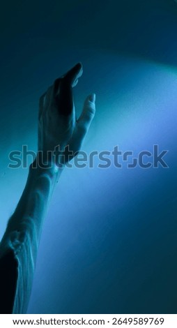 The image captures a hand elegantly reaching out towards a soothing blue light background. This intriguing interplay of light and shadow invites contemplation and imagination.