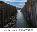 The image captures the hexagonal basalt columns of Studlagil Canyon with a river flowing through. The contrast between dark stone and green water is striking.