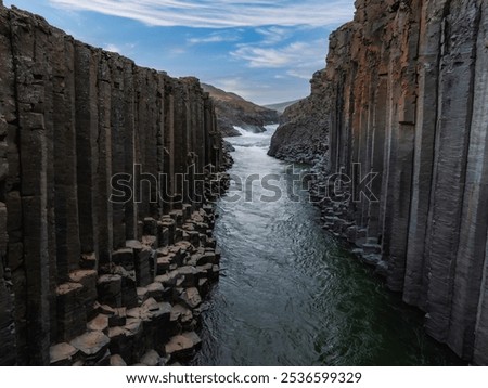 The image captures the hexagonal basalt columns of Studlagil Canyon with a river flowing through. The contrast between dark stone and green water is striking.