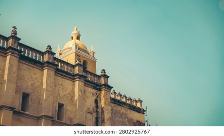 The image captures a low-angle, architectural perspective of a colonial-style church, highlighting its weathered, historic character against a clear, expansive sky. In the foreground, the solid mass of the church wall rises upward, displaying a textured, pale yellow stucco surface that shows significant signs of age, including patches of exposed stone and weathered plaster. The middle ground is defined by the church's ornate roofline, which features a series of decorative stone finials along the parapet and a central, bright yellow bell tower topped with a small dome and arched openings. In the background, the vast, unblemished sky provides a vibrant teal-blue backdrop that contrasts sharply with the earthy, warm tones of the building, creating a clean, minimalist composition. The lighting is bright and even, likely mid-day, casting soft shadows that accentuate the building's structural details and aged textures without obscuring its form. The camera angle looks upward from a ground-level perspective, which imparts a sense of grandeur and historical significance to the structure. The overall mood is serene, timeless, and classic, emphasizing the architectural beauty of the historic site.