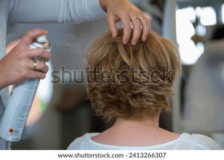 The image captures a moment of hairstyling where hands gently sculpt a short, layered haircut while applying hairspray to set the style in place. The hairstylist's hands are adorned with a wedding