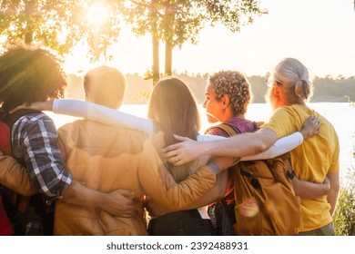 The image captures a poignant moment of four friends, seen from behind, embracing as they look out over a lake bathed in the soft light of the setting sun. The sun's rays filter through the trees