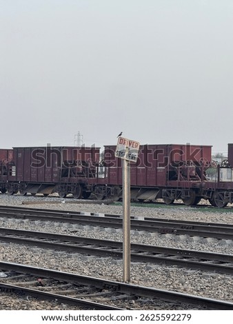 The image captures a railway yard with multiple red freight train cars aligned on parallel tracks. In the foreground, a weathered sign reads “DRIVER STOP HERE,” tilted slightly to one side. A small bi