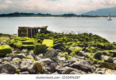 The image captures a rocky shoreline covered in vibrant green moss,with remnants of an old concrete structure. In the background,a calm sea extends towards distant mountains, under a partly cloudy sky - Powered by Shutterstock