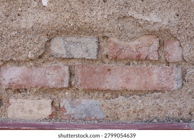 The image captures the rough surface of an aging concrete wall, where years of natural weathering and erosion have gradually worn away the outer cement layer. Tiny pebbles embedded within the concrete are exposed, creating a rich and varied texture. In some areas, the deteriorated surface reveals the red bricks hidden beneath, offering a glimpse into the structure's construction and history.
