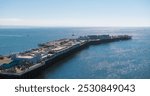 The image captures Santa Cruz Wharf with buildings and shops, surrounded by boats in the Pacific Ocean under a clear blue sky in Santa Cruz, California.