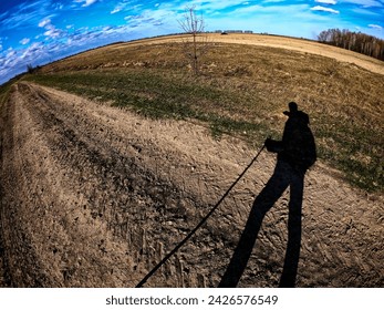 The image captures the shadow of an individual holding a stick, standing before an open field with a lone tree and distant woods.