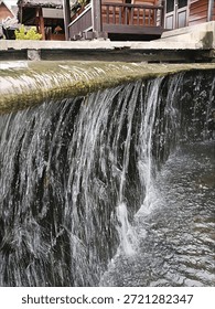 The image captures a small, cascading waterfall. Water flows over a ledge, creating a curtain-like effect as it falls into a pool or stream below. The scene is framed by a building or structure in the background.