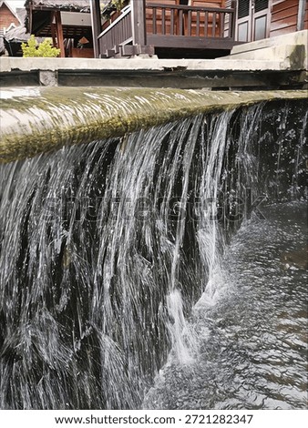 The image captures a small, cascading waterfall. Water flows over a ledge, creating a curtain-like effect as it falls into a pool or stream below. The scene is framed by a building or structure in the background.