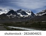 The image captures a spectacular panoramic view of the majestic Columbia Icefield peaks, highlighting Mount Andromeda and Mount Athabasca in Alberta, Canada. Large glaciers and brilliant white snowfields heavily blanket the rugged, dark limestone crags and steep alpine ridges. The lower mountain slopes drop into a barren, rocky valley basin with a rushing meltwater river under a wispy blue sky.