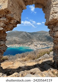 The image captures a stunning vista of a Malvasia coastal town in Greece framed by a weathered stone arch, showcasing a blend of natural beauty and historical architecture.