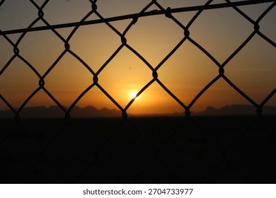 The image captures a sunset viewed through a chain-link fence. 