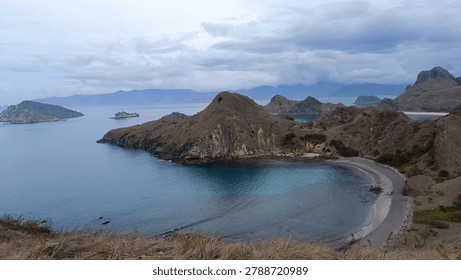 The image captures a sweeping, elevated viewpoint of Padar Island's rugged volcanic landscape in East Nusa Tenggara, Indonesia. The scene centers on a beautifully curved, crescent-shaped bay where gradient shades of deep and turquoise-blue water gently lap against a dark sand shoreline. Arid, rolling brown hills covered in dry grass and sparse vegetation sculpt the dramatic coastline, extending into prominent rocky headlands and smaller, scattered islets fading toward the horizon. Overhead, a thick blanket of dramatic, overcast clouds casts a moody and serene atmosphere across the entire landscape, muting the tropical sunlight but emphasizing the raw, prehistoric beauty of the Komodo National Park region.