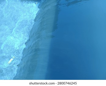 The image captures a top-down view of a small waterfall or weir barrier, dividing the water into two distinct textures. The right side features a smooth, calm, and deep blue water surface with subtle reflections and ripples. As the water flows over the edge to the left, it dramatically transforms into a turbulent, rushing stream filled with white foam, frothy bubbles, and rapid motion, creating a striking contrast between tranquility and dynamic energy.