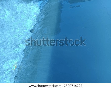 The image captures a top-down view of a small waterfall or weir barrier, dividing the water into two distinct textures. The right side features a smooth, calm, and deep blue water surface with subtle reflections and ripples. As the water flows over the edge to the left, it dramatically transforms into a turbulent, rushing stream filled with white foam, frothy bubbles, and rapid motion, creating a striking contrast between tranquility and dynamic energy.