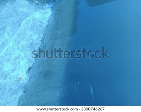The image captures a top-down view of a small waterfall or weir barrier, dividing the water into two distinct textures. The right side features a smooth, calm, and deep blue water surface with subtle reflections and ripples. As the water flows over the edge to the left, it dramatically transforms into a turbulent, rushing stream filled with white foam, frothy bubbles, and rapid motion, creating a striking contrast between tranquility and dynamic energy.