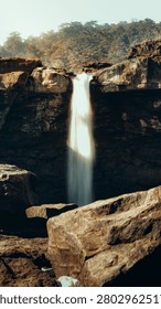 The image captures a vertical, eye-level perspective of a dramatic waterfall plunging over a rugged, rocky cliffside. In the foreground, large, jagged boulders with textured, weathered surfaces dominate the bottom of the frame, grounding the composition. The middle ground features a narrow, powerful stream of white water falling vertically into a deep, shadowed pool below, framed by dark, layered rock formations that show clear geological strata. The background transitions to a brighter, sunlit area atop the cliff, where a dense, slightly hazy forest canopy sits against a pale, clear sky. The lighting is high-contrast, with the sun casting a warm, golden glow on the top of the cliff and foreground rocks, while the area behind the waterfall remains in deep, cool shadow, emphasizing the luminosity and motion of the water. The overall colour palette is earthy, defined by deep browns, ochres, and charcoal greys of the rock, contrasted with the stark, brilliant white of the falling water and the soft green of the trees above. The mood is majestic, serene, and raw, highlighting the enduring power of nature through the sharp interplay of light, shadow, and geological form.
