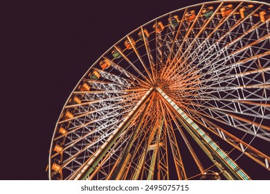 The image captures a vibrant Ferris wheel illuminated against the night sky. Viewed from below, its colorful gondolas and intricate lighting create a dramatic, festive atmosphere, inviting excitement. - Powered by Shutterstock