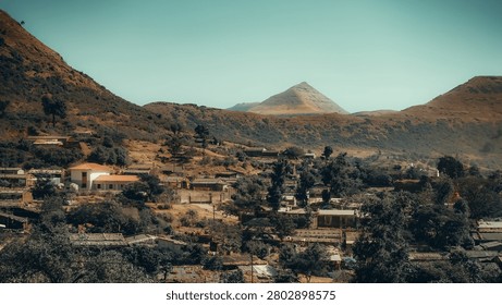 The image captures a wide, slightly elevated landscape perspective of a remote rural village nestled within a rugged, mountainous environment. In the background, a clear, pale teal sky spans above a range of rolling, rust-coloured mountains, the most striking of which is a central peak with a distinct, sharply angled, pyramidal shape. The middle ground is dominated by the sprawling settlement built directly into the uneven, dry, and rocky terrain. The vernacular architecture primarily consists of low-slung, earthy-toned structures with flat or gently pitched roofs constructed from corrugated metal and rustic materials, allowing them to blend organically with the surrounding brown and ochre earth. Contrasting sharply with these muted tones is a prominent, bright white building with a vibrant terracotta-coloured pitched roof situated on the left side of the village. The foreground features a dense, textured layering of these rustic rooftops interspersed with heavy clusters of dark, olive-green trees and shrubs that punctuate the entire settlement. Natural, bright daylight illuminates the scene from a high angle, casting clear shadows that emphasize the varied textures of the rough earth, dense foliage, and building materials, ultimately evoking a serene, isolated, and ruggedly beautiful mood.