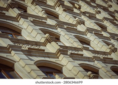 The image captures a worm's eye view of a building facade covered in pale beige stone blocks with repeating patterns of arched windows. Each window is framed by a darker brown wooden frame, and the stone work above and below features decorative elements including horizontal stepped blocks, small spherical finials, and carved floral or geometric motifs. This image is suitable for architects, designers, or real estate professionals showcasing building construction materials, facade designs, or historic architectural elements.