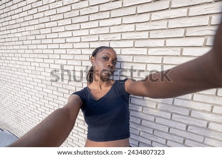 The image captures a young African woman taking a selfie with a wide-angle effect, extending her arm towards the camera. Her pose is relaxed yet expressive, with a slight tilt of her head and a calm