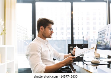 Bild eines kaukasischen Geschäftsmanns, der eine Brille trägt, die beim Arbeiten am Computer im Büro eine Zwischenablage mit Papier hält