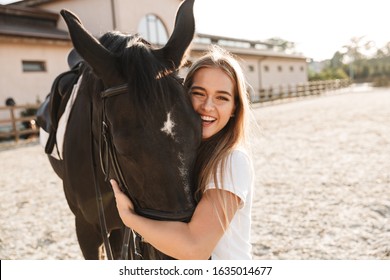 Image of caucasian cheerful positive young blonde beautiful woman with horse in countryside outdoors