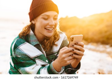Image of caucasian joyful woman using smartphone and laughing while sitting at seaside - Shutterstock ID 1617269749