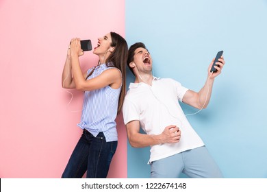 Image of caucasian man and woman wearing earphones singing while listening to music on smartphones isolated over colorful background