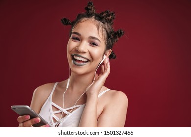 Image Of Caucasian Punk Girl With Bizarre Hairstyle And Dark Lipstick Laughing While Using Smartphone With Earphones Isolated Over Red Background