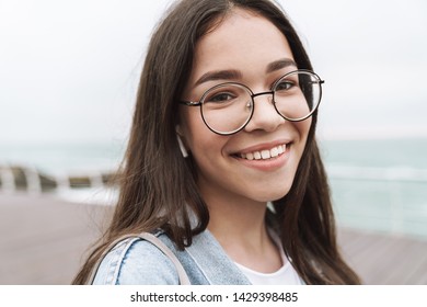 Image of caucasian teenage girl wearing earpods and eyeglasses laughing while walking along wooden pier by seaside - Powered by Shutterstock