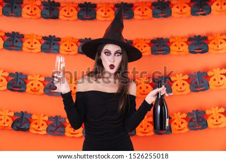 Image of caucasian witch girl in black halloween costume holding champagne bottle and glass isolated over orange pumpkin wall