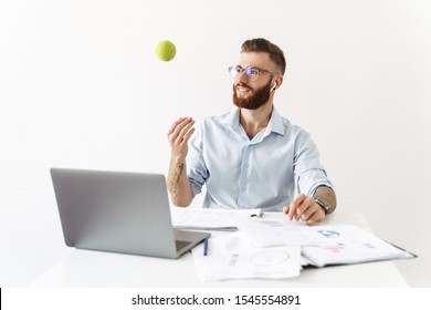 Image of caucasian young man wearing eyeglasses and earbuds playing with tennis ball while working in office - Powered by Shutterstock