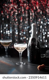 Image of celebration. The champaign bottle splashing next to which are two glasses of champaign. The background of bokeh with colors red and silver. Dark grey stone table.