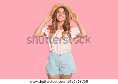 Image of charming adorable European woman touching her straw hat with both hands, looking aside, wearing blue shorts and blouse, being in high spirits, having fair hair. People and emotions concept.