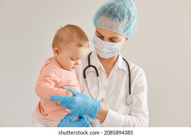 Image Of Charming Baby And Doctor Pediatrician Wearing Medical Cap, Surgical Mask, Gown And Rubber Gloves, Woman Standing With Toddler Girl In Her Arms.