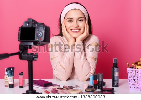Image of charming young brunette woman advertises new beauty products, makes video her blog. Beauty vlogger sits smilling in front of camera with happy facial expression. Translation of tutorial video