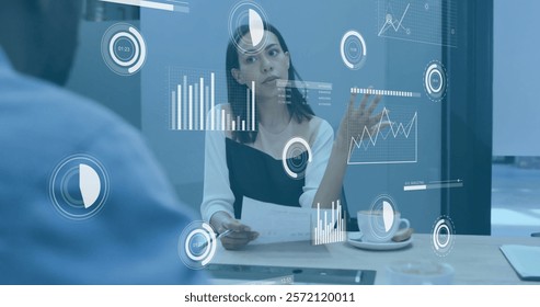Image of charts processing data over biracial businesswoman talking at casual office meeting. Business, network, digital interface and global communication, digitally generated image.