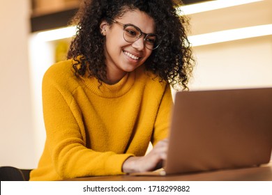 Image of cheerful african american woman in eyeglasses using laptop while sitting at living room