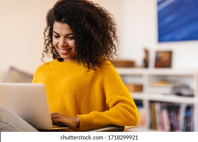 Image of cheerful african american woman using laptop while sitting on chair in living room - Shutterstock ID 1718299921