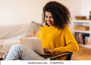 Image of cheerful african american woman using laptop while sitting on chair in living room - Shutterstock ID 1718299924