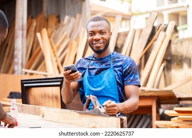 image of cheerful african man with a working tool and a smart phone- young black entrepreneur in a workshop 