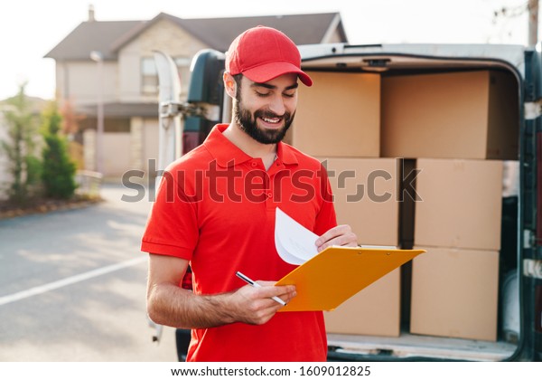 Image of\
cheerful delivery man in red uniform holding clipboard and writing\
near car with parcel boxes\
outdoors