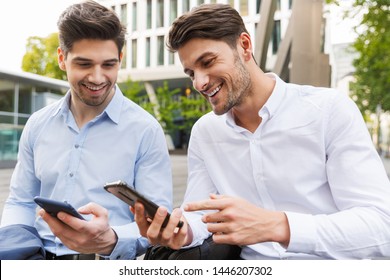 Image Of A Cheerful Happy Young Two Friends Colleagues Business Men Sitting Near Business Center Talking With Each Other Using Mobile Phones.